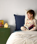 Young child sitting on a bed reading a book, with a glowing yellow bedtime lamp shaped like a smiling character placed on a green bedside table.