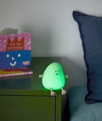 Green glowing bedtime lamp shaped like a smiling character with small arms and legs, sitting on a green bedside table next to a children’s birthday book.