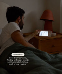 Man sitting in bed using the ergoPouch Drift Home Baby Monitor and Smart TOG Guide to check on the his child