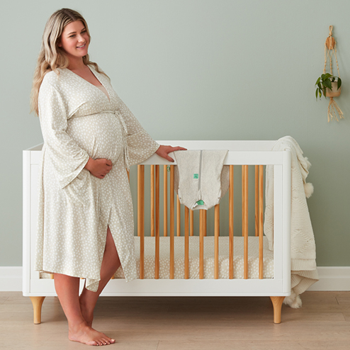 Pregnant woman standing next to crib, planning what to pack in her baby hospital bag, wearing ergoPouch Matchy Matchy Robe