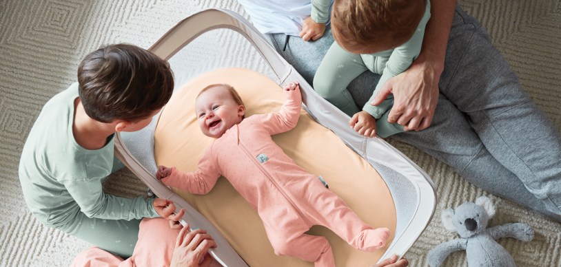  Baby laying in ergoPouch Sleep Easy Portable Crib, smiling, looking up at older siblings